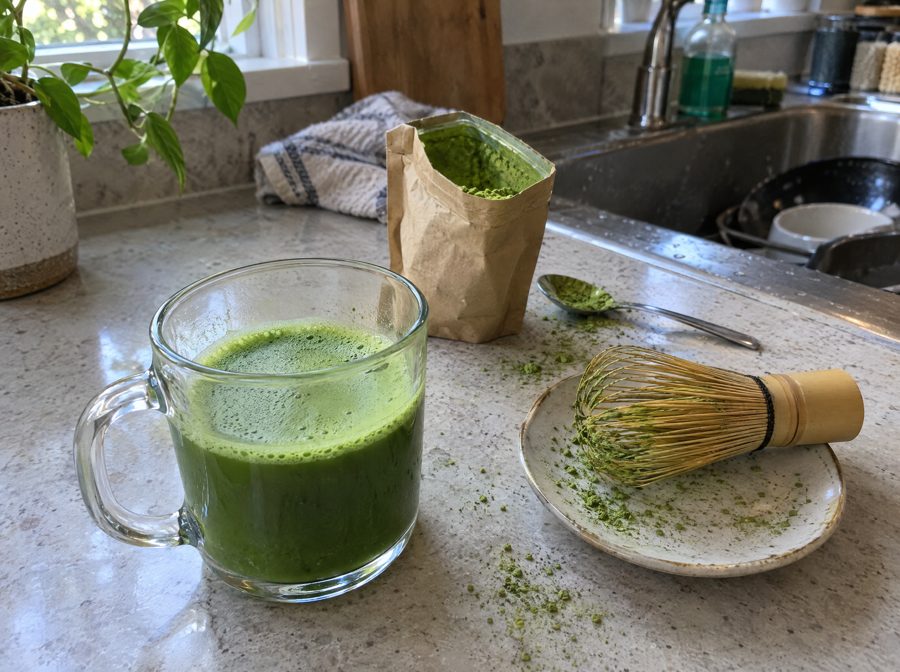 A homemade glass mug of matcha with a bamboo whisk and matcha powder on a kitchen counter